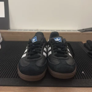Adidas Samba svart/vit sneakers - Klassiska Adidas Samba sneakers i svart skinn med vita detaljer och de ikoniska tre ränderna på sidorna. Skorna har brun gummisula, svart snörning och blå Adidas-logga på plösen. Perfekt för dig som gillar retrostil och streetwear.( Äkta morsan jobbar där)