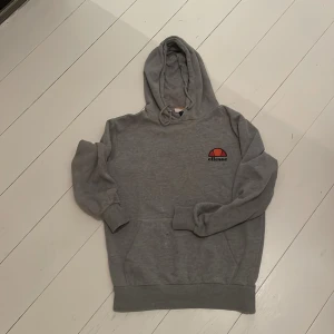 Grå hoodie från Ellesse med logga - Grå hoodie från Ellesse med klassisk logga i orange och rött på bröstet. Tröjan har huva med snörning, magficka och ribbade muddar vid ärmslut och nederkant. Perfekt för chill dagar eller när du vill ha en sportig look.