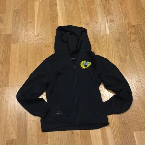 Svart Hammarby hoodie från Craft - Svart hoodie från Craft med Hammarby IF-logga på bröstet. Tröjan har dragkedja framtill, huva och två fickor. Perfekt för dig som vill visa vilket lag du hejar på. Skön och sportig stil med långa ärmar.