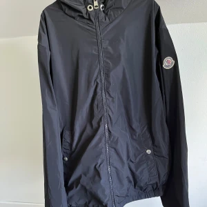 Svart vindjacka från Moncler - Svart vindjacka från Moncler med huva och dragkedja framtill. Jackan har två fickor med tryckknappar och Moncler-logga på ärmen. Lätt och smidig modell i polyester, perfekt för blåsiga dagar. Snygg detalj med röd och vit rand på huvan.