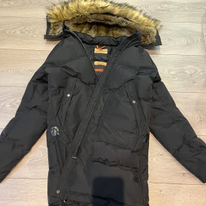 Svart lång dunjacka Parajumpers - Lång svart dunjacka från Parajumpers med stor huva och snygg fuskpäls runt kanten. Jackan har sned dragkedja, stora fickor med tryckknappar och klassisk gul detalj vid axeln. Perfekt för kalla vinterdagar och riktigt varm.