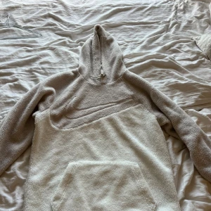 Beige fleece hoodie från Nike - Mysig beige hoodie från Nike i fleece med stor broderad swoosh på bröstet. Tröjan har huva med snörning och en stor magficka. Perfekt för kalla dagar när du vill vara både bekväm och snygg.