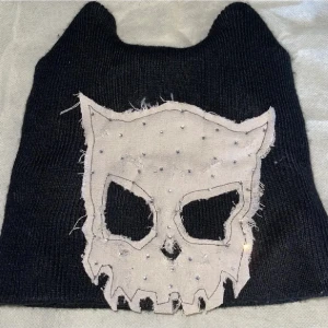 Svart mössa med döskalle och pärlor - Cool svart stickad mössa med spetsiga hörn och ett stort vitt döskallemotiv framtill. Döskallen är dekorerad med små pärlor och har en rå, fransig kant för extra edge. Perfekt för dig som gillar unik streetstyle.