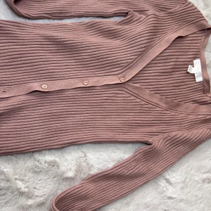 Ribbad rosa kofta från H&M - Superfin ribbad kofta i dammigt rosa från H&M. Koftan har v-ringning, långa ärmar och knäppning framtill med matchande knappar. Perfekt att slänga över en topp eller bära som den är. Materialet är mjukt och stretchigt för en bekväm passform.