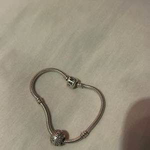 Pandora silverarmband med berlocker - Snyggt silverfärgat armband från Pandora med två berlocker, varav en är dekorerad med glittriga stenar och den andra har Pandora-loggan. Armbandet har klassisk tvinnad design och passar perfekt för att bygga på med fler berlocker.