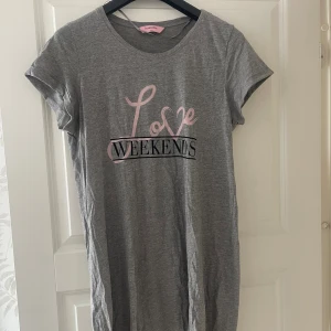Grå t-shirtklänning med tryck - Grå kort t-shirtklänning, pyjamansklänning med texten 'Love Weekends' i rosa och svart framtill. Klänningen har rund halsringning och korta ärmar. Perfekt för en avslappnad stil och gjord i mjuk bomull.