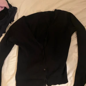 Svart kofta med knappar - En klassisk svart omsydd kofta med knäppning framtill och långa ärmar. Enkel och stilren modell som passar till det mesta. Koftan är i mjukt material och har en normal passform. Perfekt att slänga över en topp eller klänning.