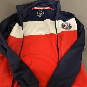 PSG vindjacka i rött, vitt och blått - Snygg PSG vindjacka i rött, vitt och marinblått med klubbens logga på bröstet och stor PARIS-text på ryggen. Jackan har dragkedja framtill och är tillverkad i polyester. Perfekt för dig som vill visa vilket lag du hejar på.