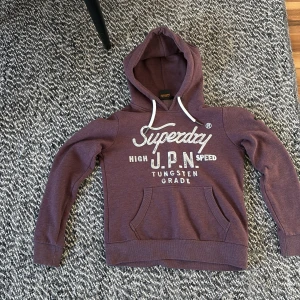 Vinröd hoodie från Superdry - Säljer en vinröd hoodie från Superdry med vit logga och text på bröstet. Tröjan har huva med vita snören. Perfekt för dig som gillar archive och vill ha en snygg och bekväm tröja. Tröjan är liten i storleken och sitter mellan s och xs.