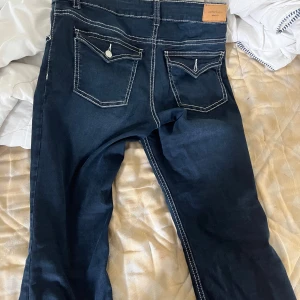 Mörkblå jeans med kontrastsömmar - Snygga mörkblå jeans från Perfect Jeans med vita kontrastsömmar och unika bakfickor med lock och knappdetalj. Klassisk femficksmodell med dragkedja och knapp framtill. Perfekta för dig som gillar detaljer och stilren look.