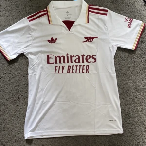 Vit Arsenal fotbollströja Adidas M - Snygg vit Arsenal fotbollströja från Adidas med vinröda detaljer och klubbens kanon-logga på bröstet. Tröjan har klassisk krage, röda ränder på axlarna och guldfärgade inslag på ärmsluten. Tillverkad i lätt och ventilerande Aeroready-material.