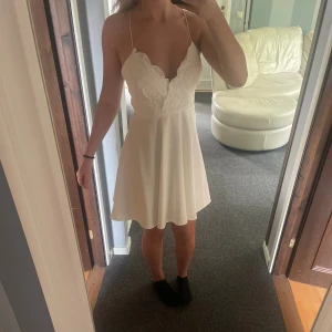 Vit spetsklänning med tunna axelband - Supersöt vit kortklänning med spetsdetaljer upptill och tunna korsade axelband i ryggen. Klänningen har en v-ringad omlottliknande överdel och en lätt utsvängd kjol. Perfekt för sommarens alla tillfällen eller studenten! Stretchig så funkar för s-m. Aldrig använd.