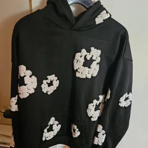 Svart hoodie från Denim Tears med vita blommor - Cool svart hoodie från Denim Tears med stora vita blommor tryckta över hela tröjan. Oversized passform, huva och känguruficka framtill. Perfekt statement-plagg för dig som vill sticka ut. Mjuk insida och riktigt skön att bära.
