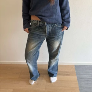 Levi's 501 blå baggy jeans - Säljer ett par klassiska Levi's 501 jeans i blå tvätt med slitningar och ljusa partier. Jeansen har låg midja, raka ben och en baggy passform. Materialet är robust denim och byxorna har fem fickor samt råa kanter nertill för en avslappnad vibe.