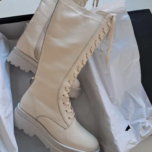Beiga höga boots i skinn med snörning (Zign) - Snygga höga boots från Zign i beige skinn med grov platt sula, varmt foder och snörning längs hela framsidan. Skorna har dragkedja på insidan för enkel påtagning och en spetsig tå som ger en cool vibe. Perfekta för dig som vill sticka ut med stilrena detaljer. 