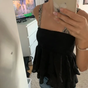 Svart volangig linnetopp - Supersöt svart linnetopp med volanger och breda axelband i beige spets. Smockad över bysten för en bekväm passform och flowy nedtill. Perfekt att styla med jeans eller kjol för en trendig look.