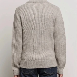 Beige stickad kofta från Tiger of Sweden - Snygg beige stickad kofta från Tiger of Sweden med ribbad struktur och hög krage. Koftan har hel dragkedja framtill och är långärmad, perfekt för lager-på-lager. Materialet är mjukt och värmande, och passar dig som gillar stilrena plagg med premiumkänsla.