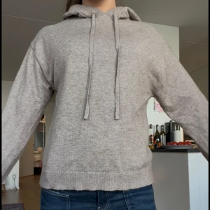 Beige stickad hoodie med huva - Mysig beige stickad hoodie med huva och snörning. Tröjan har lång ärm och ribbade muddar vid ärmslut och nederkant. Perfekt för dig som gillar en enkel och stilren look.