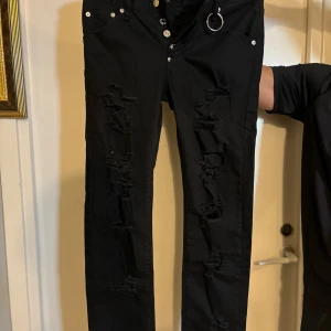 Svarta Dsquared2 jeans med slitningar - Svarta jeans från Dsquared2 med tight passform och snygga slitningar längs benen. Byxorna har silverfärgade knappar, femficksmodell och en cool metallring vid ena fickan. Materialet är bomull med stretch för extra komfort. Perfekta för dig som gillar en edgy streetstil.
