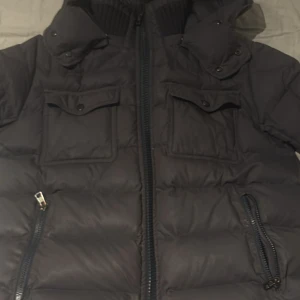 Svart Moncler pufferjacka med huva - Svart pufferjacka från Moncler med två stora bröstfickor, dragkedja framtill och huva. Jackan har Moncler-logga på ärmen och coolt serietidningsfoder på insidan. Perfekt för kalla dagar och riktigt snygg streetstil. Pris kan diskuteras