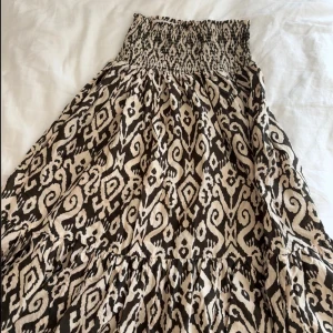 Mönstrad långkjol från Zara  - Supersnygg långkjol från Zara i beige och svart med ett unikt grafiskt mönster. Kjolen har smock upptill och faller snyggt i flera volanger. Perfekt för dig som gillar boho-vibbar och vill sticka ut med coola detaljer.