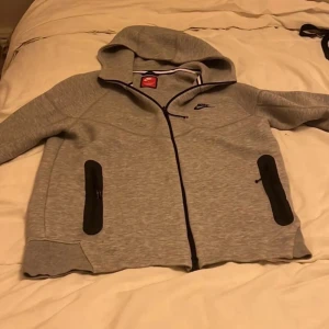 Grå hoodie från Nike med dragkedja - Grå hoodie från Nike med svart logga på bröstet, hel dragkedja och två svarta fickor med dragkedja. Tröjan har huva och är långärmad, perfekt för chill eller träning. Materialet är mjukt och känns som polyester. Snygg och enkel design.