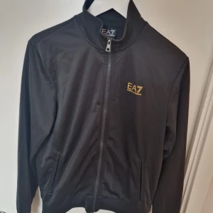 Svart vindjacka EA7 Emporio Armani - Svart tracksuit från EA7 Emporio Armani med guld logga på bröstet och snygga guldfärgade detaljer på ryggen. Jackan har dragkedja framtill, ståkrage och två sidofickor. Perfekt för dig som gillar stilren och sportig design. Dessutom får du med de stiliga byxorna med loggan på baksidan av byxorna 