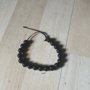 Svart pärlarmband med justerbar knytning - Snyggt armband med svarta glittriga pärlor på elastisk tråd och justerbar knytning. Perfekt accessoar för att ge din outfit lite extra edge. Pärlorna har en cool glansig finish som fångar ljuset på ett stilrent sätt.