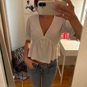 Vit blus med v-ringning och volang - Supersöt vit blus med djup v-ringning och volang nedtill. Blusen har korta ärmar med lätt puff och är gjord i ett ribbat material med diskreta ränder. ( obs se diefekt )