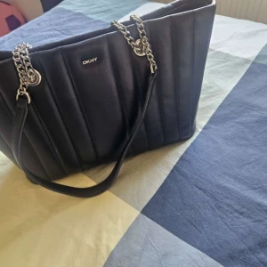 Svart quiltad handväska från DKNY - Snygg svart handväska från DKNY med quiltad design och silverfärgade kedjehandtag. Väskan är rymlig och har en stilren look som passar perfekt till din vardagsstil. Tillverkad i skinn med vertikala sömmar för extra detalj.