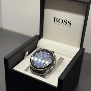 Blå kronografklocka från Hugo Boss - Snygg kronografklocka från Hugo Boss med blå urtavla och silvrigt armband i rostfritt stål. Klockan har tydliga index, tre subdials och robust länkarmband. Kommer i originalask med märkets logga. Perfekt accessoar för dig som gillar stilren design.