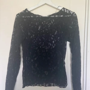 Svart spetsblus från Pieces - Svart långärmad blus från Pieces i genombruten spets med rund halsringning. Toppens spetsmönster ger en snygg och trendig look. 