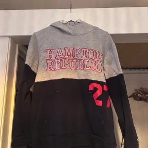 Grå och marinblå hoodie Hampton Republic - Snygg hoodie från Hampton Republic i grått och marinblått med stor logga och siffran 27 i rött på ryggen. Tröjan har huva med tryck, lång ärm och känguruficka framtill. Perfekt för en chill och sportig stil.