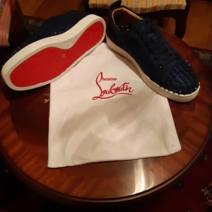 Christian Louboutin sneakers med nitar - Mörkblå sneakers från Christian Louboutin med klassisk röd sula och coola nitar på tån. Skorna har snörning, rund tå och är tillverkade i mocka med läderdetaljer på insidan. Perfekta för dig som vill sticka ut med en lyxig och edgy stil.