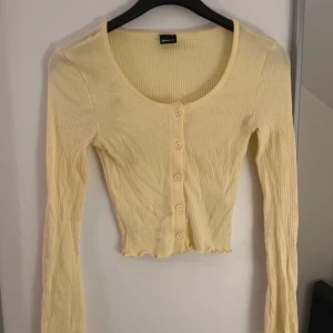 Gul ribbad croppad topp med knappar - Söt ljusgul croppad topp från ginatricot med långa ärmar och ribbad struktur. Toppen har knappar framtill och en lätt vågig kant nertill för en extra detalj. Perfekt för dig som gillar en enkel men trendig look.