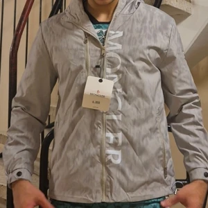 Grå vindjacka från Moncler med huva - En helt ny moncler jacka. Jag säljer den då den är för liten för mig. Den passar en liten M                                                                                Snygg grå vindjacka från Moncler med stor logga framtill, huva med dragsko och dragkedjor på fickorna. Jackan har långa ärmar, diskret Moncler-märke på ärmen och är tillverkad i lätt polyester. Perfekt för dig som gillar stilren och sportig look.