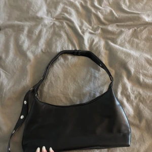 Svart axelväska i skinn med nitar - Säljer en svart axelväska i skinn med coola silvriga nitar på axelremmen och dragkedja upptill. Väskan har en stilren, avlång form och justerbar rem. Perfekt för dig som vill ha en enkel men edgy accessoar till din outfit.