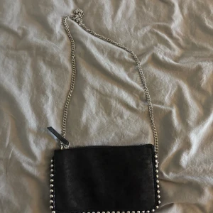 Svart axelväska med silverkedja - Snygg svart axelväska i mocka med coola silverfärgade nitar runt kanterna och en lång kedjerem. Väskan har dragkedja upptill och flera fack inuti för att hålla ordning på dina grejer. Perfekt storlek för det viktigaste och ger en edgy vibe till din outfit.