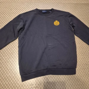 Mörkblå sweatshirt från GANT - Mörkblå sweatshirt från GANT med rund hals och gul broderad logga på bröstet. Tröjan har långa ärmar och ribbade muddar vid ärmslut och nederkant. Perfekt för en stilren och klassisk look.
