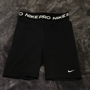 Nike Pro svarta träningstights-shorts - Svarta Nike Pro shorts i tight modell med elastisk midja där det står 'NIKE PRO' i vitt. Perfekta för träning eller sport. Swoosh-logga framtill på benet. Gjorda i stretchigt syntetmaterial för maximal rörelsefrihet.