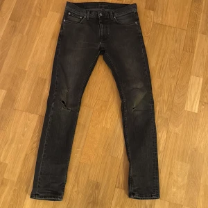 Svarta jeans från Nudie Jeans W30 L34 - Svarta jeans från Nudie Jeans med skinny passform och klassisk femficksdesign. Jeansen har slitningar och hål på knäna för en cool, avslappnad look. Materialet är denim i bomull och de har svarta detaljer på bakfickorna. Storlek W30 L34. Ny pris 1699kr säljer då dom ej passar längre 