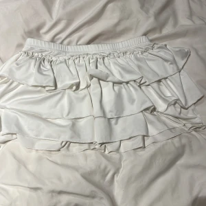 Vit volangkjol med rosett - Supersöt vit kortkjol med flera lager volanger och en stor rosett framtill. Kjolen har elastisk midja och det är inbyggda shorts 