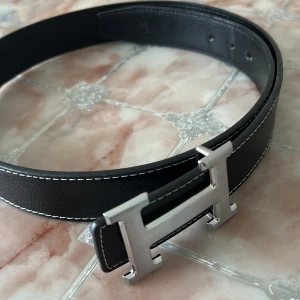 Svart H  skärp med H-spänne - Stilrent svart skärp från  H med ikoniskt H-spänne i silverfärgad metall. Bältet är gjort i präglat skinn och har vita kontrastsömmar längs kanterna för en snygg detalj. Perfekt accessoar för att lyfta din outfit.