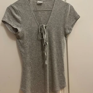 Grå ribbad topp med snörning Gina Tricot - Grå ribbad topp från Gina Tricot i storlek S. Modellen har korta ärmar, v-ringning och snygg snörning framtill. Mjuk och stretchig i materialet. 