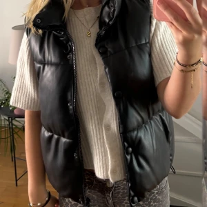 Svart dunväst i skinnimitation från Zara - Säljer en svart dunväst från Zara i skinnimitation. Västen har hög krage, dragkedja och tryckknappar framtill. Perfekt att slänga över en stickad tröja för en snygg look🤩 Storlek Xs