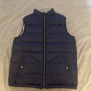Mörkblå dunväst från Ralph Lauren - Säljer nu min snygga mörkblåa dunväst från Ralph Lauren med diskret logga på bröstet. Västen har två fickor framtill med knappstängning och dragkedja hela vägen upp. Perfekt för lager-på-lager och håller dig varm under kyliga dagar. Köpt ny- 1700 på rea. Priset är ej hugget i sten👍🏼