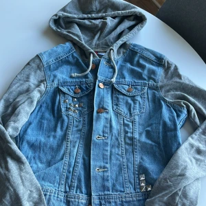 Jeansjacka med grå luvärm från Divided - Snygg jeansjacka från Divided med grå luvärm och huva i sweatshirtmaterial. Jackan har coola nitar på bröstfickan och nedtill, samt knappar i kopparfärg. Perfekt för dig som gillar streetstyle och vill sticka ut lite extra.