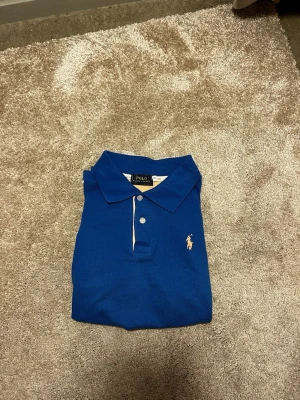 Blå pikétröja från Polo Ralph Lauren - Snygg blå pikétröja från Polo Ralph Lauren i storlek S. Klassisk krage, två knappar och ikonisk gul broderad logga på bröstet. På bilderna ser det ut som att de är färgen gul på logo och insidan men det är ljusrosa. Tröjan är gjord i mjuk bomull som är skön mot huden och passar perfekt till jeans eller shorts. Priset ej hugget i sten hör av er vid frågor eller funderingar! 