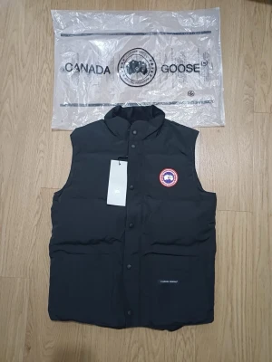 Svart dunväst från Canada Goose - Svart dunväst från Canada Goose med klassisk ståkrage och tryckknappar framtill. Västen har ett broderat rött och vitt logomärke på bröstet och två stora fickor med Canada Goose-tryck. Materialet är slitstark polyester och dunfyllning för extra värme.