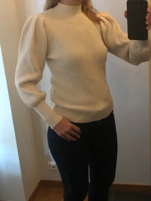 Beige ribbstickad tröja med puffärm - Säljer en beige stickad tröja med ribbad struktur och hög hals. Tröjan har långa puffärmar och breda muddar vid ärmslut. Tröjan är liten i storleken och mer som en M.